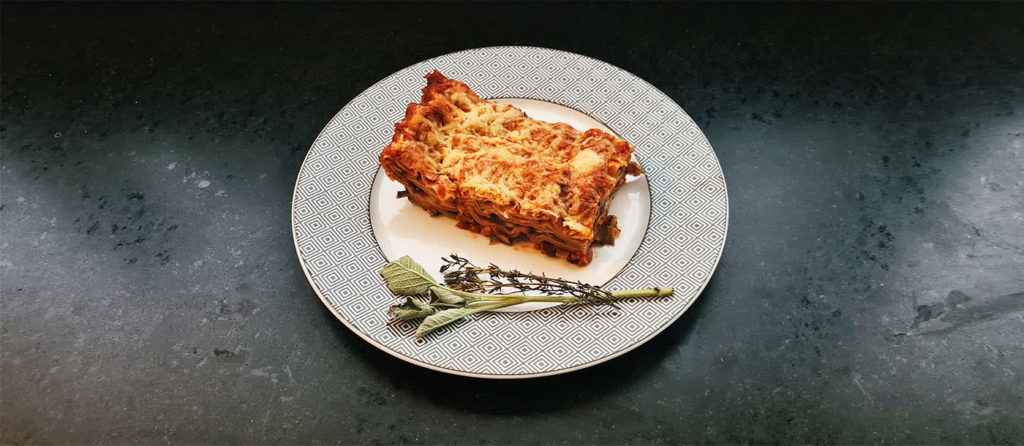 Ein quadratisches Stück Lasagne auf einem Teller mit frischem Thymian und Salbei